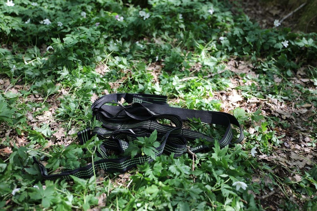 Svarta vävda hängmattaremmar med gröna sömmar liggande på marken omgiven av grön vegetation och vita blommor