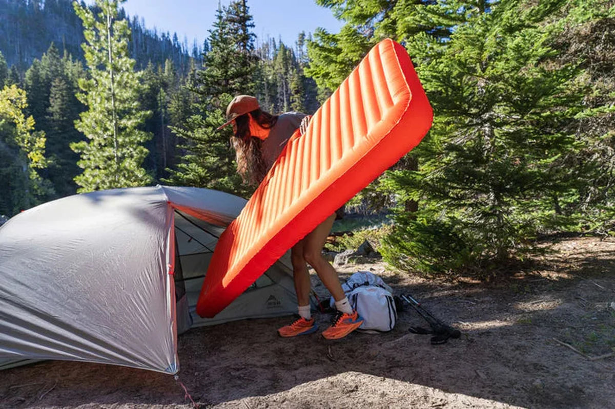 Kvinna med orange NeoLoft liggunderlag vid campingtält i skogsmiljö