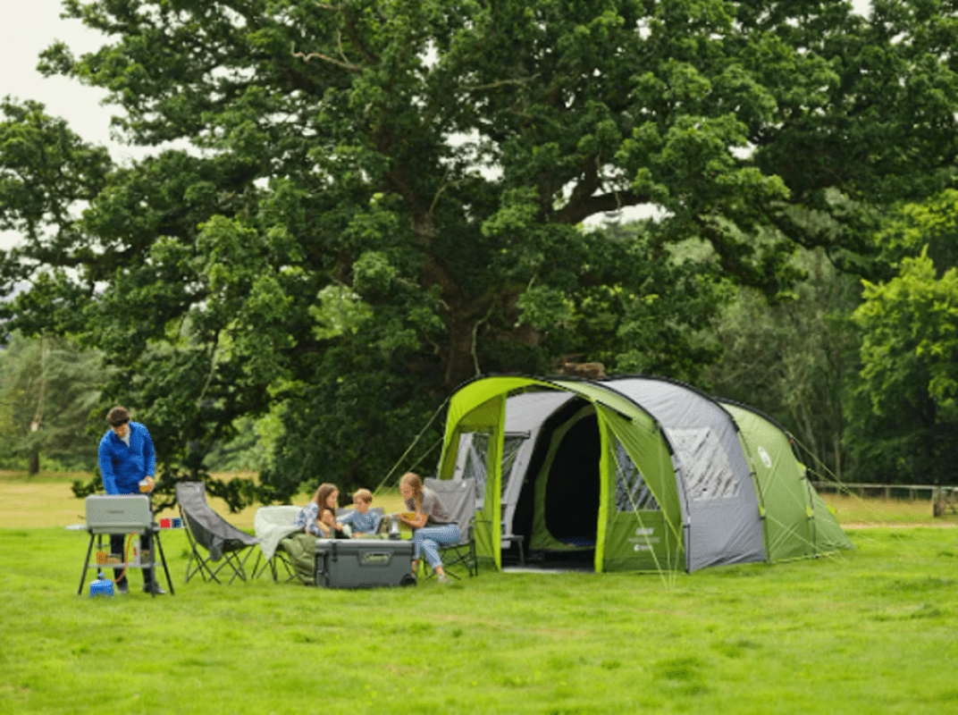 Familj vid grill utanför öppen veranda på Coleman Rocky Mountain 5 campingtält under träd