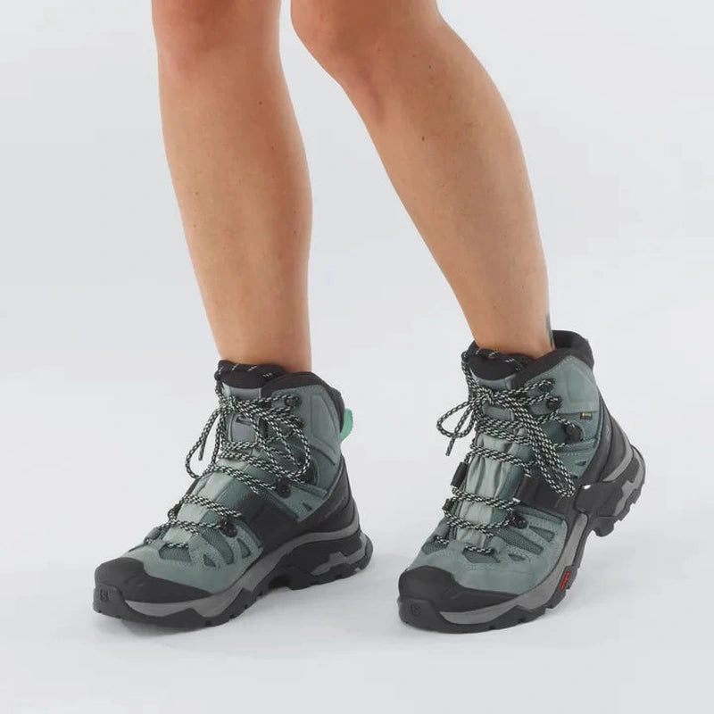 Modell som bär grågröna Salomon Quest 4 GTX med teal dragkedja mot vit bakgrund